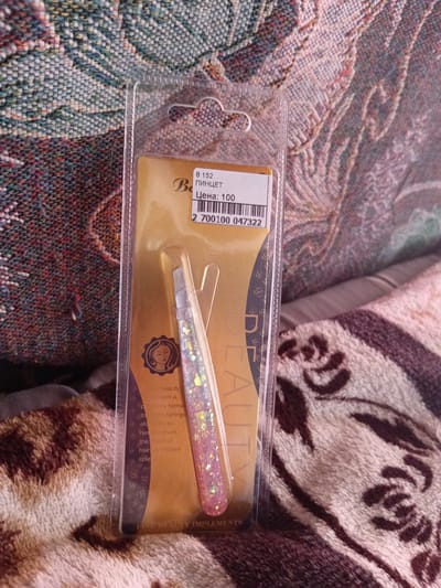 The image shows a pair of glitter-covered tweezers in its original retail packaging. The packaging is clear plastic with a gold backing card. The backing card has text printed in Russian, including "8 152 Пинцет" (8 152 Tweezers) and "Цена: 100" (Price: 100). Below this text is a barcode and the number "2 700100 047322". The tweezers are pink and iridescent with glitter. The packaging is lying on a patterned textile surface that appears to be a blanket or upholstery. The background is blurred but suggests a domestic environment. The specific location in Monastirische, Russia, cannot be determined from the image alone, as there are no identifiable landmarks or context clues to suggest a city.