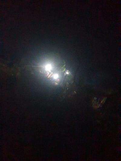 A dark outdoor scene with bright streetlights illuminating a path or road. The lights cast a glow, suggesting a wet or reflective surface. Trees are vaguely visible. The background is shrouded in deep shadows, making it difficult to discern details. The scene may be a forest road or a street in a natural setting at night.
