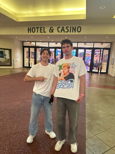 The image shows two males standing in front of the entrance to the Hollywood Seminole Hotel & Casino. The male in the foreground is wearing a white t-shirt and blue jeans, and is smiling. The male behind him is wearing a white t-shirt with a graphic design on the front and grey pants. The location is the Hollywood Seminole Reservation, United States of America.