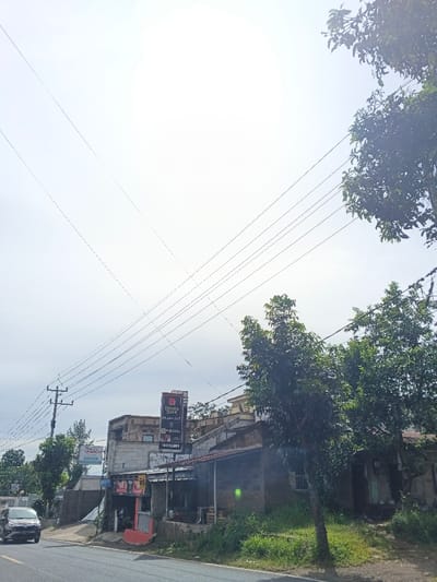 A street scene features a small, weathered building with a red sign and a larger sign above it. Power lines crisscross overhead against a bright sky. Green trees frame the right side of the image. A car drives along the asphalt road in the lower-left corner. The scene conveys a sense of rural or suburban simplicity.
