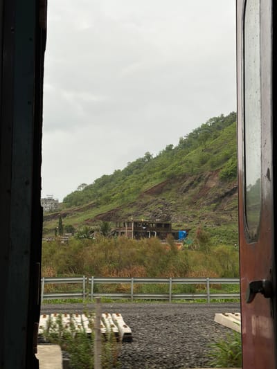 The image, taken from inside a train carriage looking out through an open doorway, displays a cloudy sky over a lush green, heavily vegetated hillside. The hillside shows signs of a landslide with exposed reddish-brown earth and rocks. At the base of the hill, partially obscured by trees and vegetation, are several unfinished or abandoned construction sites with exposed concrete structures. In the mid-ground, a metal guardrail runs parallel to a railway track, which is laid on a bed of dark gravel. In the foreground, several concrete railway sleepers are visible on the gravel. The doorway frames the view on both the left and right sides, with the left side showing a dark, weathered wooden frame and the right side a reddish-brown door with a handle. The overall scene suggests a rural or semi-rural setting near the outskirts of Vasai-Virar, India, possibly during or after monsoon season, given the greenery and the landslide.