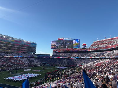 A bustling stadium filled with cheering fans clad in red, white, and blue, creating a patriotic visual display. A large screen displays a live broadcast. The field hosts a large flag and a group of performers. The stadium, branded as "Levi's Stadium," also features a prominent Pepsi logo. The sky above is clear and bright. The overall scene captures the excitement and grandeur of a major sporting event.