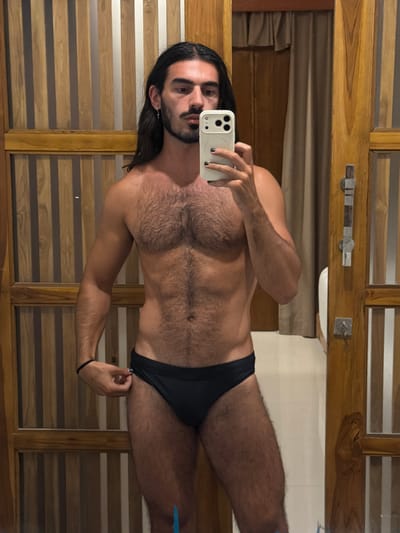 A person is taking a mirror selfie in a bathroom. The person has long dark hair and a beard, and is wearing black swim briefs. They are holding a white smartphone in their left hand. The background appears to be a wooden wall with horizontal slats and a beige curtain. A silver towel rack is visible on the wall. The lighting suggests it is daytime, possibly indoors. The overall impression is a casual, personal moment.