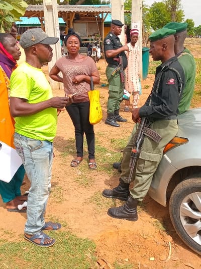The image shows a group of people in Ungwan Tanko, Nigeria, located at latitude: 10.422, longitude: 7.49. There are five individuals in the scene. Two of the individuals are police officers in uniform, one standing and one crouched, holding rifles. The standing police officer is wearing a dark green uniform with a black beret. The crouched police officer is wearing a dark green uniform with a green beret. The officers are positioned beside a silver car.
There is a woman in a light brown shirt and dark pants standing near the police officers, and two women standing near the front of the image. One of the women is wearing a light blue shirt and dark pants, and the other is wearing a brightly colored dress. The woman in the brown shirt is looking at the officers, and the woman in the dress is speaking to the man standing between her and the officers. This man is wearing a green t-shirt and blue jeans. A second man is standing near the crouched officer, facing the other individuals in the image. He is wearing a cream-colored shirt with a white, blue, and brown cap. He is looking at the officers. All the individuals in the scene are standing on a dirt ground. There is a building to the left side of the image. Trees and vegetation are visible in the background.