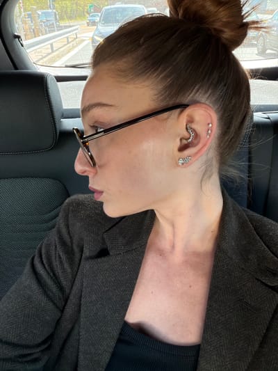 A woman is photographed from the side, appearing to be seated in a vehicle. She has her brown hair pulled back in a bun and is wearing glasses. Several piercings are visible in her ear, including a helix piercing with three small studs, a rook piercing with a curved, jeweled piece, and a lobe piercing with a floral-shaped earring. She is wearing a dark blazer over a dark shirt. The background shows a blurry view of a road and vehicles, suggesting an outdoor setting. Given the prompt constraints including the location of Serravalle Scrivia, Italy, it can be inferred that the photo was taken inside a vehicle in Serravalle Scrivia.