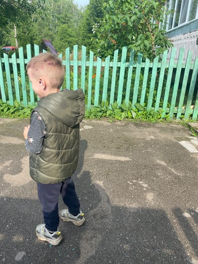 An outdoor daytime scene in Ihnatpil, Ukraine, features a young child positioned with their back mostly to the camera, standing on a dark grey asphalt surface. The child is wearing a dark green hooded puffer vest over a grey long-sleeved shirt with multi-colored star and rainbow patterns, dark blue trousers, and dirty grey and white athletic sneakers with chunky soles and yellow accents. Circular reddish-purple markings are visible on the child's scalp and behind their left ear.
Behind the child, a vibrant green wooden picket fence runs horizontally across the mid-ground. Beyond the fence, dense green vegetation, including trees and bushes, is present. In the far left background, a partially visible red object, resembling a vehicle component, and a weathered wooden structure are discernible amidst the foliage. To the far right, a portion of a grey building with vertical window panes and textured siding is visible.
The asphalt ground is uneven, exhibiting patches of dampness or stains, and is traversed by distinct sunlight and shadow patterns. Small green plants grow along the base of the green fence. A metal grate is visible on the ground in the lower right corner of the frame.