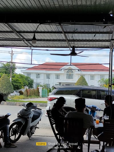 This image depicts a scene at an outdoor cafe in Ben Cau, Vietnam, viewed from under a corrugated metal roof. The foreground is dominated by silhouettes of people seated at tables, some with drinks. To the left, a white scooter is parked. In the mid-ground, a white SUV is parked on a street lined with trees and bushes. Beyond the street, a large, white building with a red tiled roof stands prominently, featuring multiple windows and a central arched window with decorative glass. The sky is partly cloudy, suggesting it's daytime. A ceiling fan is visible overhead. The timestamp "2026.06.24 09:40" and the text "realme Shot by zsm_travel" are overlaid on the image. The overall atmosphere appears casual and relaxed, consistent with a cafe setting.