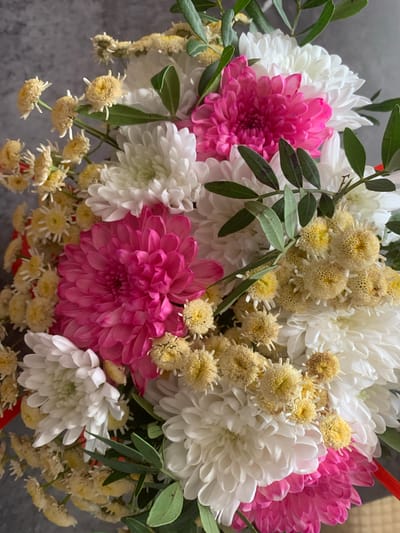 A bouquet of flowers, including pink and white chrysanthemums and small yellow button-like flowers, is displayed against a grey, textured background. The flowers appear to be arranged loosely, with some greenery interspersed. The lighting suggests an indoor setting. There is no indication of a specific location in Starodub, Russia.