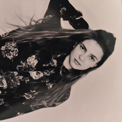 The image is a black and white portrait of a female subject, angled approximately 45 degrees to the lower right. The subject has long, dark hair, styled to appear windswept, cascading over her left shoulder and chest. She is wearing a dark-colored garment with a floral pattern, featuring lighter-toned flowers. Her left arm is raised, with her hand positioned near the top of her head, partially obscured by her hair. Her face is turned towards the camera, with a direct gaze. Her mouth is closed, and her expression is neutral. The background is a plain, light-colored surface, suggesting a studio or controlled environment. There are no other discernible objects or subjects in the frame. The visual evidence presented does not provide information to determine a specific location within the city of Dubovka, Kazakhstan, or any other geographic location.