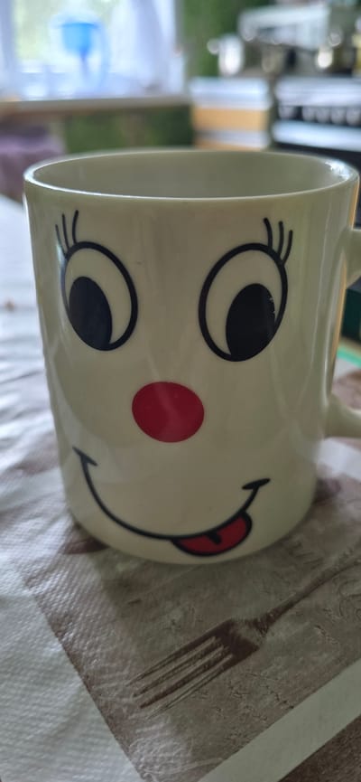 A close-up, eye-level shot captures a ceramic mug with a cartoonish face design. The mug is off-white and features large, black-outlined eyes with blue irises and prominent black eyelashes. A bright red circle serves as a nose. Below the nose, a black line forms a wide smile with a red tongue sticking out. The mug sits on a tablecloth with a checkered pattern of brown and white stripes, on which a faint image of a fork is visible. In the background, a blurred window with a blue object and a kitchen counter with containers can be seen. The lighting is bright, suggesting an indoor setting during the daytime. The location is specified as Dankov, Russia, though no specific city landmarks or features confirm this.