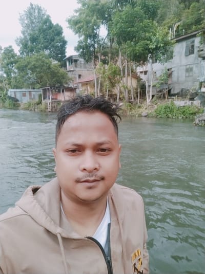 A man is taking a selfie in front of a body of water. He has dark hair and a mustache. He is wearing a tan hooded jacket over a white shirt. The water is a murky green and appears to be a river or stream. In the background, there are several buildings and trees on a hillside. One building looks like it is made of concrete and has several windows. Another building is made of corrugated metal and wood. The overall scene suggests a rural or semi-urban environment in a tropical or subtropical region. The weather appears to be overcast, as the lighting is diffused and there are no strong shadows. There is no clear indication of the time of day, but the overcast conditions might suggest it is mid-morning or afternoon. The man's expression is neutral, and he is looking directly at the camera. There is a patch on the man's jacket that has some text and graphics, but it is difficult to read clearly. The location context provided is Blang Bebangka, Indonesia.