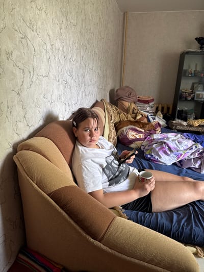 The image shows a young person lying on their back on a sofa, with their head resting on the armrest. They are wearing a white t-shirt and black shorts. In their right hand, they are holding a white mug with a strawberry pattern, and in their left hand, they are holding a smartphone. The person has dark hair and is looking towards the camera with a neutral expression.
The sofa is upholstered in a light brown fabric. Behind the sofa, there is a cluttered shelf with various items, including some framed pictures, what appear to be toiletries, and some fabrics. The room is dimly lit, suggesting it might be evening or nighttime. The wall behind the shelf has a textured, light-colored wallpaper. The floor is covered with a light-colored, patterned linoleum. A dark blue blanket is visible on the sofa. The overall impression is of a casual, indoor setting, possibly a living room or bedroom. The location context provided is Valday, Russia.