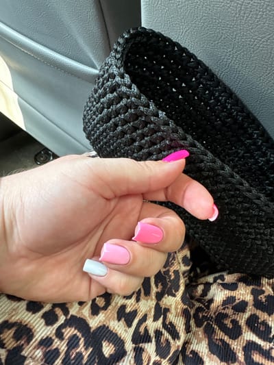 The image shows a close-up of a person's hand with brightly colored fingernails, set against a backdrop of textured materials. The hand is the primary subject, with its nails painted in a mix of neon pink, light pink, and a pale grey. The texture of the skin on the hand is visible, with fine lines and creases.
The background consists of three distinct elements. To the left, there is a dark, textured material that appears to be crocheted or knitted, with a woven pattern. In the center and right, a fabric with a prominent leopard print pattern is visible, featuring the characteristic spots and swirls in shades of brown, black, and beige. There is also a hint of a grey, textured surface, possibly upholstery, in the upper left corner, suggesting the setting might be inside a vehicle or a room with leather seating.
The lighting suggests it is daytime, with a natural light source casting subtle shadows. There are no visible signs of a specific event or activity, other than the presentation of the hand and its manicure. The overall impression is one of personal style and grooming. No text is visible in the image.
