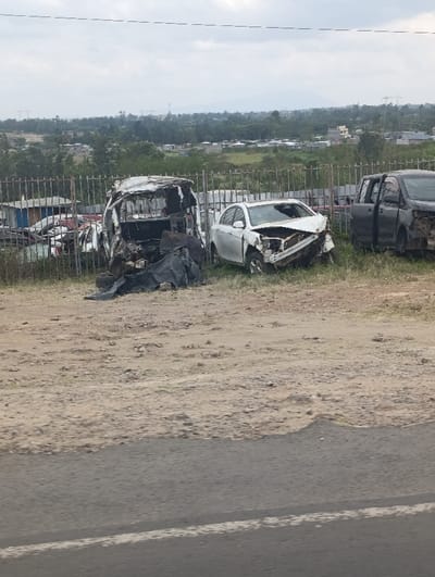 The image presents a static scene of three heavily damaged vehicles in an outdoor impound or salvage lot, located in Matungulu west ward, Kenya. In the foreground, a section of asphalt road with a single white line transitions to a strip of dry, light-brown dirt and small rocks. A metal fence separates this area from the vehicle storage. Behind the fence, from left to right, are the primary subjects: a white van or mini-bus, severely burnt and structurally compromised with an exposed, blackened interior; a white sedan, likely a Toyota Corolla, exhibiting extensive front-end collision damage; and a dark-colored SUV or pickup truck, featuring significant side damage with its passenger door ajar. Additional vehicle wreckage and debris are visible in the background of this section. The environment beyond the fence comprises scattered trees, numerous distant low-rise buildings, and indistinct mountains or hills under an overcast sky. Overhead power lines are visible in the upper portion of the frame. No individuals are present, and no active events are depicted.