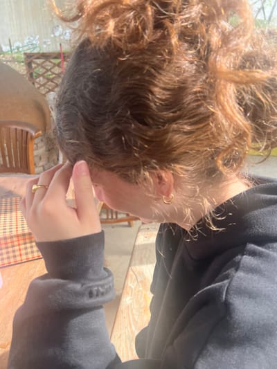 The image displays a close-up, side profile view of an individual, likely female, observed from the right, situated in an outdoor or semi-outdoor setting during daylight. The individual is facing towards the left of the frame.
The subject has wavy, light brown hair gathered in an updraft, possibly a messy bun or ponytail, with some loose strands framing the right ear and nape of the neck. They are wearing a dark-colored, long-sleeved hooded garment, consistent with a sweatshirt. A single, small, gold-colored hoop earring is visible in the right earlobe. A plain gold-colored band ring is worn on the ring finger of the right hand. The right hand is raised, with fingers gently pressed against the temple or side of the head, partially obscuring the right side of the face.
In the foreground, a light-colored wooden table or surface is present. A reddish-brown and beige checkered pattern, possibly a placemat or a printed design on the table, is visible to the left of the subject's hand.
The background features elements of an outdoor environment. A portion of a curved, dome-like structure, constructed of stone or brick with a lighter, possibly stucco or clay finish, is visible behind the subject. Adjacent to this structure, a wooden bench or chair with a slatted back is partially discernible. Further behind, a light-colored wooden trellis or fence is present, with hints of green foliage beyond it.
The scene is brightly illuminated by natural sunlight, casting highlights on the individual's hair and the wooden surfaces.
While the location is specified as Hof Ashkelon Regional Council, Israel, the visual elements within the image do not provide any specific identifying features or landmarks to forensically confirm this geographical detail. The scene depicts a generic outdoor leisure or dining area.