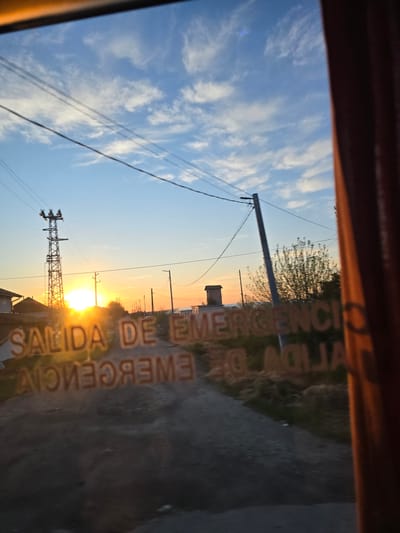 This image, presented rotated 90 degrees clockwise, captures a sunrise or sunset scene from inside a vehicle looking out a window. The bright, golden sun is prominently visible on the lower right (east), casting a warm glow across the horizon. The sky above is a clear blue with scattered, wispy white clouds, transitioning to soft orange and yellow hues closer to the sun.
In the middle ground, several utility poles and a larger, lattice-style power transmission tower stand against the colorful sky, with numerous power lines stretching across the frame. Beyond these, distant trees and a small, cuboid structure, possibly a water tower or technical building, are silhouetted. The foreground consists of a dark, indistinct ground surface, likely a road or field, and a dark, blurred section at the top edge, indicating the interior of the viewing vehicle.
Reflected prominently on the glass surface through which the photo was taken are the mirrored and inverted Spanish words "SALIDA DE EMERGENCIA" (Emergency Exit), indicating the view is from an emergency exit window. The overall atmosphere suggests a peaceful moment at dawn or dusk in Glojene, Bulgaria, characterized by infrastructure and natural beauty.