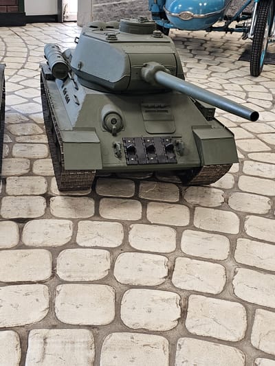 A high-angle shot shows a military tank on a stone pavement. The tank has a long, cylindrical gun barrel pointing diagonally upwards and to the left. The tank is green. Next to the tank is a small part of a vintage motorcycle in blue. The background is the white stone pavement. The location is not discernible from the given image.