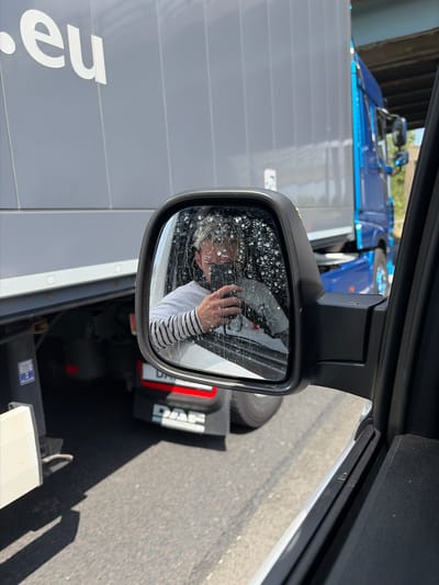 The image shows a person taking a selfie in a side-view mirror of a truck. The person, with short blonde hair and wearing a striped shirt, is holding up a phone to capture the reflection. The mirror is dirty with water spots and streaks, obscuring some of the reflection.
The setting appears to be a roadside or industrial area, with a large grey truck trailer in the foreground and a blue truck in the background. The trailer has white text that is partially visible, reading ".eu". The blue truck has "DAF" branding visible on its side. The ground is asphalt, suggesting a road or parking lot. Sunlight indicates it is daytime, likely midday given the brightness. The overall mood is casual, as indicated by the selfie. No specific emotions are discernible beyond the act of taking a photograph. The location context of Rochdale, United Kingdom is noted, but not directly evident from the visual elements within the image itself.