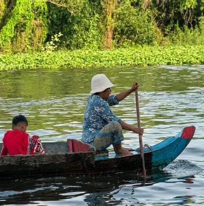 A woman wearing a white hat and a floral blue shirt is rowing a small wooden boat. She is sitting in the boat, holding an oar and paddling through the water. A child wearing a red shirt is sitting in the front of the boat, facing away from the camera. The boat is on a body of water, with lush green vegetation lining the background. The scene appears to be in a rural or natural setting, possibly a river or canal. The water is calm and reflects the greenery. The lighting suggests it is daytime. The location is not explicitly shown in the image, but the style of the boat, clothing, and environment are consistent with rural areas in Southeast Asia. Given the context of Soutr Nikom, Cambodia, this image likely depicts a scene from that region, where traditional boats and water transportation are common.