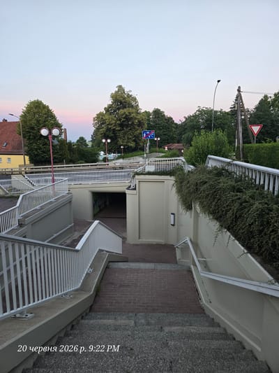 The image depicts an underpass in the city of Bannewitz, Germany, with a pedestrian staircase leading down into it. The underpass is lit by artificial light, and graffiti is visible on the walls. Above the underpass, a pedestrian bridge with white railings spans across the scene.

The surrounding environment is urban, with trees and buildings visible in the background. A traffic sign indicating a pedestrian crossing is present, along with a yield sign. The sky is a soft twilight hue, suggesting it is either dawn or dusk.

The timestamp on the image indicates it was taken on June 20, 2026, at 9:22 PM. No people are visible in the image. The overall scene is one of a quiet urban passage, possibly in a residential area.