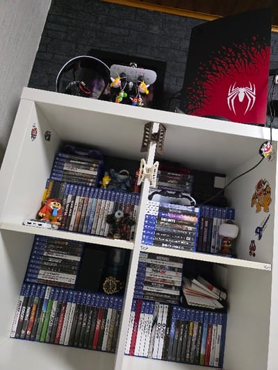 This image depicts an indoor scene within a residence in Zweibrücken, Germany, focusing on a white modular shelving unit positioned against a wall. The unit is primarily populated with gaming-related items.

The white shelving unit has at least three horizontal shelves and two vertical dividers, creating six visible cubbies and a top surface. The wall behind the unit features dark, possibly charcoal or black, faux-brick textured wallpaper or paneling. A dark wooden trim or ceiling is visible at the top edge of the frame.

On the top surface of the shelving unit, from left to right: a black over-ear gaming headset is present. To its right, a white display stand holds multiple small, colorful keychains, including characters resembling Mario and Luigi. Behind these, a black framed object is partially visible. On the far right, a black vertical gaming console, likely a PlayStation 5, stands upright. Its front panel is adorned with a custom red and black "Spider-Man" themed skin featuring a prominent white spider logo. Cables are visible connected to the console.

The cubbies of the shelving unit are organized as follows:
*   The top-left cubby contains stacks of PlayStation 4 game cases, a small white electronic device, a figurine resembling Link from "The Legend of Zelda," and a dark, armored figurine.
*   The top-right cubby holds stacks of PlayStation 4 game cases, with a white PlayStation DualShock 4 controller resting on top of one stack. A Funko Pop! figurine with a white shirt is positioned on the right.
*   The middle-left cubby is filled with PlayStation 4 game cases. A dark-colored smartphone lies flat on top of some cases, next to a gold-colored wristwatch with a black bezel and a dark cylindrical object.
*   The middle-right cubby contains more PlayStation 4 game cases, with a stack of horizontally oriented white-spined cases or books on the right.
*   The bottom-left and bottom-right cubbies are filled with standing PlayStation 4 game cases.

Several stickers are affixed to the white shelving unit: a small red and white sticker on the upper left vertical divider, a red and white circular sticker near the top of the right vertical divider, and a collection of stickers on the far right side of the unit, including a large yellow and brown character resembling Bowser, a blue number "9", and smaller brown mushroom-shaped icons. Two white, multi-jointed plastic or 3D-printed objects are suspended from the underside of the top shelf, positioned centrally.