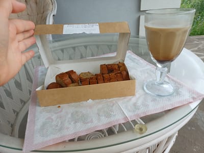 In the city of Chernovskiy, Russia, a hand is seen reaching into a cardboard box containing sliced banana bread. The box is open and placed on a white, patterned paper napkin with a pink border, which rests on a transparent glass table. Next to the box, a clear glass with a stem holds a light brown beverage, likely coffee with milk or cream. The table itself is set on a white, woven outdoor furniture base. In the background, a portion of an upholstered chair is visible, along with a grey wall and greenery suggesting an outdoor or semi-outdoor setting. The lighting suggests daytime.