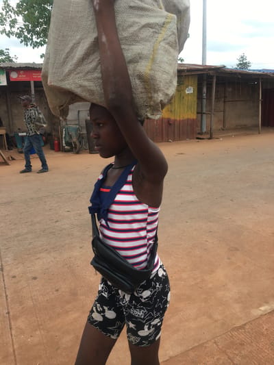 The image captures a vibrant street scene in Isieke, Nigeria, during bright daylight. The main subject is a young individual with dark skin, positioned in the foreground, carrying a large, light-colored woven sack on their head. They are wearing a red, white, and blue striped tank top, black shorts patterned with white Mickey Mouse motifs, and a black bag slung across their body. Their expression is neutral and focused.
The setting appears to be a busy thoroughfare, possibly a market area, characterized by a light brown, possibly concrete, road. In the midground and background, several simple buildings and stalls line the street. These structures are constructed from corrugated metal sheets, some painted yellow and red-brown, with concrete or rough-hewn walls. One building has a blue tarp roof.
Other individuals are visible in the background, including a man in a plaid shirt and blue jeans standing near one of the structures. Various objects are scattered around, such as a utility pole, green trees and foliage, a red fire extinguisher, a wheelbarrow, and metal pipes.
Visible text includes a prominent red "Airtel" sign and partially visible white text below it that reads "REGISTER SIM CARD" with the number "891" also present. A white sign with black, illegible text is affixed to a yellow corrugated door. The activity centers around daily life and commerce, with people moving and goods being transported. The weather is clear, and the overall impression is one of an active, functional community street.