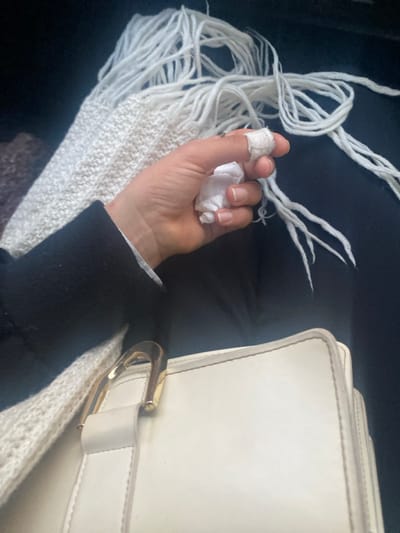 The image captures a close-up, downward perspective of a person's upper legs, lap, and right hand within an interior setting in the city of Bembla, Tunisia. The visible right hand has its thumb wrapped in white bandage material and is holding crumpled white tissue paper. The person wears a dark-colored long sleeve, with a white cuff partially visible, and dark trousers or a skirt. A white, coarsely knitted or woven textile with long fringes, possibly a scarf or blanket, is draped across the lap, primarily on the left. To the right, a light-colored (off-white or cream) structured handbag with a prominent gold-toned metal clasp or buckle rests on the lap. The background is dark and out of focus.