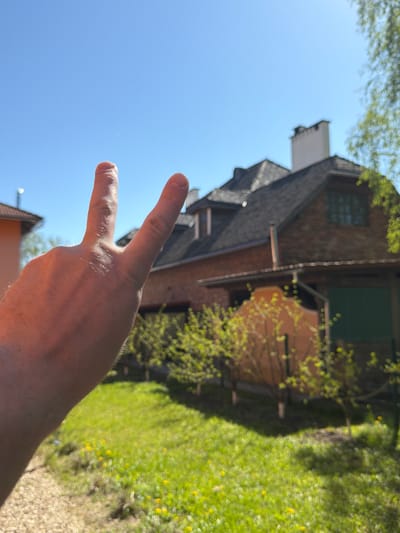 A low-angle, slightly rotated outdoor shot shows a person's hand making a peace sign against a backdrop of buildings and greenery. The hand is in the lower left quadrant of the image, with two fingers extended upwards and the rest of the hand receding towards the lower right corner. The skin tone appears light.
Behind the hand, a building with a dark gray shingle roof is partially visible. It features a brick exterior, a white chimney-like structure, and a green window on the upper level. The architectural style appears traditional.
To the right of the building, a patch of green grass with small yellow flowers is visible. A tree or bush with light green leaves is also present, creating a soft, out-of-focus background.
The sky is visible in the upper left quadrant of the image and is a clear blue, suggesting a sunny day.
There are hints of a roof of a building in the very bottom of the frame, it's colored orange.
It is not possible to confirm that the location is Тэльмінскі сельскі Савет, Belarus from the image alone.