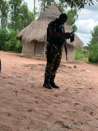 An individual in military-style camouflage fatigues, a dark tactical vest, and black combat boots stands on reddish-brown dirt ground in Sankara, Nigeria. The person, positioned facing slightly left, is holding and looking down at a small dark electronic device in their hands. An assault rifle, possibly an AK variant, is slung across the individual's front. In the background, to the left, is a traditional circular hut with a conical thatched roof and a small wooden door. Dense green foliage and trees are visible behind the hut and further in the background, under an overcast sky. The foreground and midground consist of packed earth with scattered dry leaves and small debris.