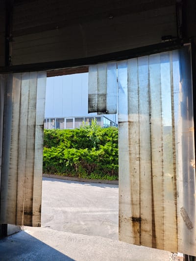 The image captures a view through a translucent, dirty strip curtain, likely at an industrial loading dock or warehouse entrance in Hofgeismar, Germany. The curtains are made of vertical plastic strips, some of which appear torn or heavily soiled with dark smudges and streaks. Sunlight streams through the curtain, casting shadows on the concrete ground in the foreground. Beyond the curtain, a paved area leads to a dense green hedge. Behind the hedge, a modern building with a light-colored facade and rows of windows is visible. The sky is a clear light blue. The overall scene suggests an outdoor industrial or commercial setting with signs of wear and tear on the entrance material.