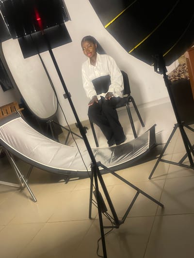 A young woman is seated on a chair in what appears to be a photo studio. She is wearing a white button-down shirt and black pants. She has dark hair pulled back and is wearing makeup. In front of her is a large, curved reflective surface, possibly a beauty dish or a curved backdrop. Behind her, to the left, is a large softbox light. To the right, another softbox light is visible. The room has a tiled floor and a white wall. The overall impression is that of a staged photoshoot. The location is described as South Ropp, Nigeria, but this cannot be visually confirmed from the image content.