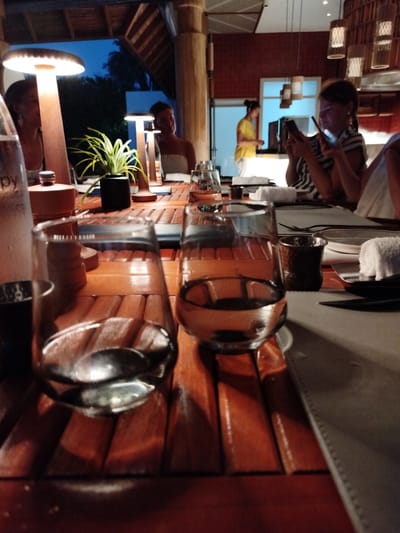 The image depicts a dimly lit, outdoor dining scene at dusk. In the foreground, two glasses of water are prominently displayed on a wooden table, with spoons resting inside. The table is set with placemats, napkins, and small metal cups. Behind the glasses, a plant in a dark pot adds a touch of greenery. Further back, several people are visible, some appearing to be dining or conversing. One woman, in the middle ground, is engrossed in her smartphone. In the background, a person in a yellow top stands near a doorway. The setting appears to be a restaurant or resort, possibly in a tropical location given the lush foliage visible outside. The ambient lighting suggests it is evening, and the overall mood is relaxed and intimate. The word "apy" is visible on a water bottle in the left foreground.