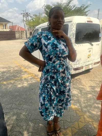 A young adult female is depicted standing outdoors in Ozuaha, Nigeria, under bright daylight. She faces slightly right of the camera but turns her head to look directly forward, displaying a slight open-mouthed smile. Her hair is styled in braids, and she wears a short-sleeved, midi-length dress featuring a V-neck and a white, teal, and dark blue/black abstract or camouflage pattern, with ruched detailing on the left side of the skirt. Her left hand rests on her hip, and her right hand is positioned near her chin. She wears dark sandals with decorative elements. Behind her is the rear of a white utility van, identified as a "HIJET" model, bearing a Nigerian license plate "NSR-39XC" and a blue wheelchair accessibility sticker. The ground is paved with light grey interlocking concrete pavers featuring yellow painted lines. In the background, a light-colored building with a dark roof and a dark perimeter wall is visible to the left, along with utility poles and wires against a bright sky. Scattered green foliage is present further in the background. Shadows are cast on the paved surface, indicating overhead sunlight.
