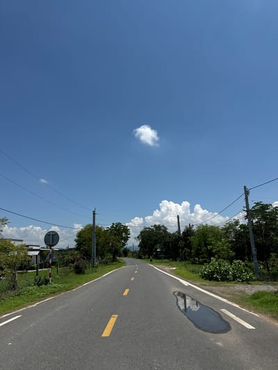 The image shows a straight, paved road with a white line on the left edge and yellow dashed lines in the center, indicating it is a two-way street. The road is surrounded by lush green grass and trees on both sides. In the foreground on the right, there is a large puddle reflecting the sky. On the left side of the road, there is a red and white striped pole with a circular sign. Further down the road, there are power lines supported by poles. The sky is a vibrant blue with a few scattered white clouds. In the distance, the road curves slightly and disappears behind a line of trees and hills. The scene appears to be in a rural or suburban area of Hồng Sơn, Vietnam, characterized by its natural landscape and infrastructure. There are no people visible in the image.