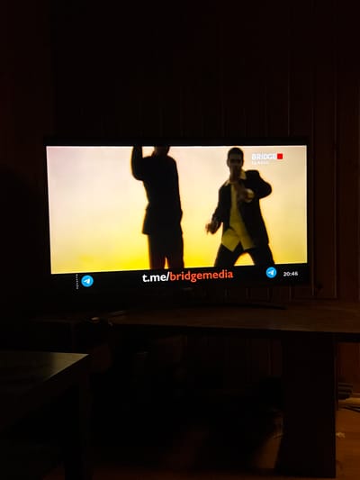 In a dimly lit room, a television screen displays a scene with two silhouetted figures against a bright yellow-orange gradient background. The figure on the left is wearing a dark jacket and appears to be raising an arm. The figure on the right is wearing a dark jacket over a yellow shirt and is in a dynamic pose. At the top right of the screen, a logo reads "BRIDGE CLASSIC". Along the bottom of the screen, from left to right, are the words "PIXAMANA", a blue Telegram icon, the text "t.me/bridgemedia", and the time "20:46". The television is placed on a dark surface, possibly a desk or table, which is mostly obscured by shadow. The background of the room consists of vertical wooden paneling. The scene is static, with no discernible activity outside of what is depicted on the television. The location is identified as Bykovo, Russia.