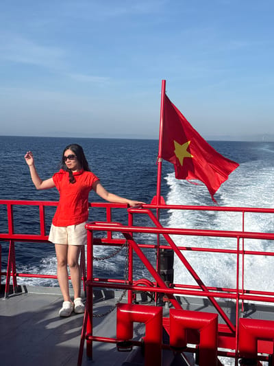 A woman stands on the deck of a boat, facing left, with her right arm extended upwards. She is wearing a red short-sleeved shirt and light-colored shorts, and sunglasses. A red flag with a yellow star, likely the Vietnamese flag, is visible on a flagpole to her right. The boat is moving through calm blue water, leaving a white wake behind it. In the distance, a hazy landmass can be seen on the horizon. The deck railing is painted red. The scene appears to be set in the open sea, not specifically within the city of Dong Son, Vietnam, though it could be near its coast.