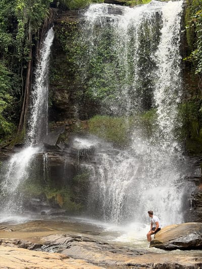 A person is sitting on a large rock at the base of a waterfall. The person is wearing a white t-shirt and black shorts. Their legs are submerged in the water. The waterfall is surrounded by lush green foliage and moss-covered rocks. The water is cascading down a rocky cliff face, creating a misty spray. The ground in the foreground is composed of large, flat rocks. The scene appears to be in a natural, outdoor environment, possibly a park or a jungle. This location is identified as being in the city of Ban Huai Takhrai, Thailand.