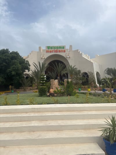 The image shows the exterior of a building with large arched entrances, likely a hotel or resort, located in Taguermess, Tunisia. The building is white with decorative stepped architectural elements. Above the central entrance, the words "Welcome Meridiana" are displayed in colorful lettering. The entrance area is surrounded by lush green grass, palm trees, and various plants. In the foreground, there are wide, light-colored concrete steps leading up towards the building. The sky is overcast with a few patches of blue visible. There are no people visible in the image. Various potted plants and some decorative elements, such as a large clay amphora, are placed around the entrance and landscaping.