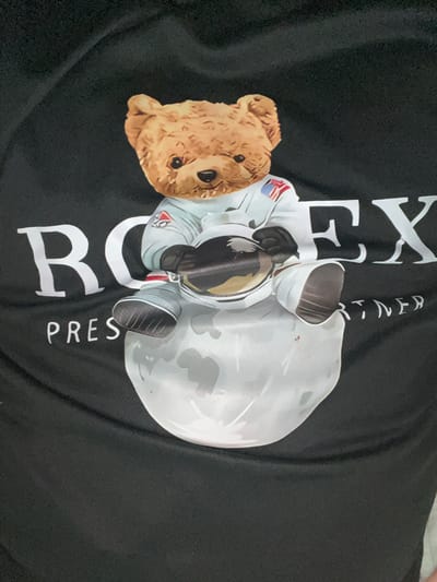 A teddy bear dressed as an astronaut is depicted sitting on a moon. The teddy bear is wearing a white astronaut suit with an American flag patch on the left shoulder and a blue and red patch on the right chest. It is holding a golden, reflective helmet. The text "RO" is visible to the left of the bear, and "EX" is visible to the right, in large white block letters. Below the "RO," the word "PRES" is also visible in white letters. The background is black fabric, likely a t-shirt, which is slightly creased. The image does not provide any information about the city of Rosenovo, Bulgaria.