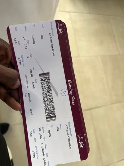 A person is holding two boarding passes for Value Jet airline with the passenger name ATTAH/ABRAHAM. The flight is from JOS to LAGOS on April 19 at 1405 (2:05 PM). The flight number is VK 253 and the class is S. The seat number is 15A and the boarding time is 1335 (1:35 PM). The location is not visible in this image, and the mention of Kwi, Nigeria is not relevant to the image contents.