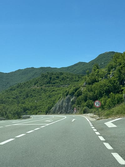 The image depicts a winding asphalt road in a mountainous area, likely in or near Očinići, Montenegro, under a clear blue sky. The road is marked with white dashed lines and solid white lines indicating lanes and directions. On the right side of the road, there is a circular speed limit sign displaying "70" in red and white, and a triangular warning sign with a graphic of a winding road. The road curves to the right, leading towards a forested mountainside covered in lush green trees. Rocky outcrops are visible on the left side of the road, and more mountains covered in dense vegetation extend into the distance. The scene appears to be a scenic view of a mountain road, possibly a route that passes through Očinići. There are no people or vehicles visible in the image, and the environment is natural and undeveloped, emphasizing the road and the surrounding landscape.