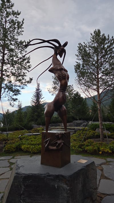 A bronze sculpture of a mythical creature stands in an outdoor park. The creature has the head and upper torso of a human-like figure with elongated, tendril-like appendages extending from its head. Its body is slender with muscular legs, and it is positioned in a dynamic, bent posture. The sculpture is mounted on a square bronze pedestal that features a relief of a reclining nude figure on one of its sides. This pedestal, in turn, rests on a large, rough-hewn dark grey stone base. The park setting includes lush green bushes, scattered rocks, and mature pine trees. In the background, a body of water can be seen on the left, with rolling hills and a cloudy sky. This sculpture is located in the city of Ozernoye (Mayskiy), Russia.