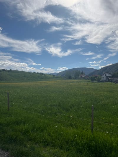 The image presents a rural landscape with a blue sky filled with streaks of white clouds dominating the left side. On the right side, a large grassy field extends towards a distant mountain range. The field appears to be dotted with small yellow flowers and is enclosed by a wire fence with wooden posts. A few houses can be seen nestled at the base of the mountains. The image seems to be rotated. There is no indication of the location being in Madzhare, Bulgaria. There are no people or animals visible in the scene.