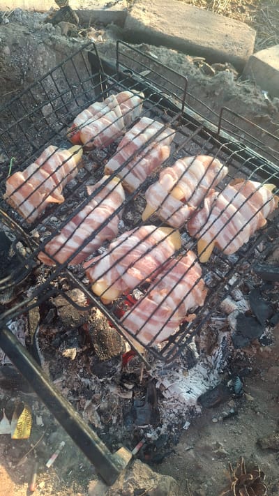 The image depicts food being grilled over coals. The food items, likely sausages or similar cylindrical items, are wrapped in strips of bacon and placed on a metal grill grate. The grill is positioned over a bed of glowing coals and ash, indicating an outdoor cooking setting. The surrounding environment appears to be rustic, with dirt, rocks, and dried vegetation visible. There are no people or animals present in the frame. The scene is consistent with a casual outdoor cooking event. The location in Навасвержанскі сельскі Савет, Belarus is not ascertainable from the visual information alone.