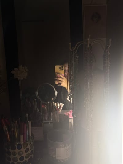 A mirror reflects a cluttered space: a person holding a phone, makeup containers, tubes, and a small round mirror. Above the mirror, jewelry hangs from a rack. A framed photo is visible on the left. In the foreground, a flower arrangement sits, partially obscured. The scene is dimly lit, with a bright light source at the top.