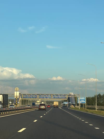 The image shows a highway in Zimnyaya Gorka, Russia, under a partly cloudy blue sky. The perspective is from the driver's seat of a vehicle moving along the highway. In the foreground, the asphalt road has white dashed lane markings. On the left side of the road, a metal barrier separates the lanes from the shoulder, where a large white truck is visible. Further down the highway, other vehicles, including cars and trucks, are traveling in the same direction.
Overhead, a pedestrian bridge crosses the highway. Blue panels are visible on the bridge's structure. Several lampposts line the right side of the highway, with trees and a fence visible beyond them. A blue road sign with white text and an arrow points to the right, indicating directions to Orenburg and Turgay. The overall scene depicts a typical highway environment in Russia during daylight.