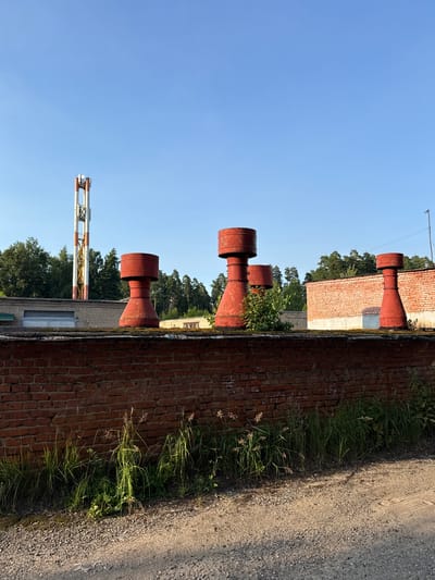 The image shows a brick wall in the foreground, with tall grass growing along its base. Behind the wall, there is a flat roof with four red, conical chimney-like structures. Further back, a long, low brick building with a large garage door is visible, and beyond that, a forest of green trees. To the left of the trees, a tall, white and red striped radio tower stands against a clear blue sky. The scene appears to be an industrial or utility area on the outskirts of the city of Солнечная Поляна Кп, Russia. There are no people or animals visible in the image.
