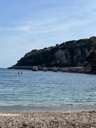 Captured in Saint-Jean-Cap-Ferrat, France, the image presents a sunlit day by the sea. A rocky cliff face, densely covered with trees, forms the backdrop, adjacent to the clear, rippling water. Two people are visible on a paddleboard in the sea. A beach of pebbles and debris fills the right foreground. The sky is light blue.
