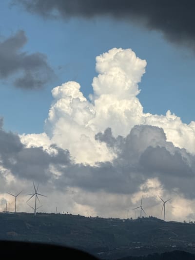 The image shows a landscape view of rolling hills with wind turbines on the crest, located in Đa Lộc, Vietnam. The sky is filled with dramatic clouds, ranging from dark, ominous storm clouds in the upper portion of the frame to bright, voluminous cumulus clouds in the center. Several wind turbines are visible on the ridge, silhouetted against the sky. The foreground features a dark, out-of-focus area, likely the interior of a vehicle or a person's obstruction. Buildings and structures are scattered across the hillsides, suggesting a populated area. The overall impression is one of a dynamic and expansive natural landscape under a turbulent sky.
